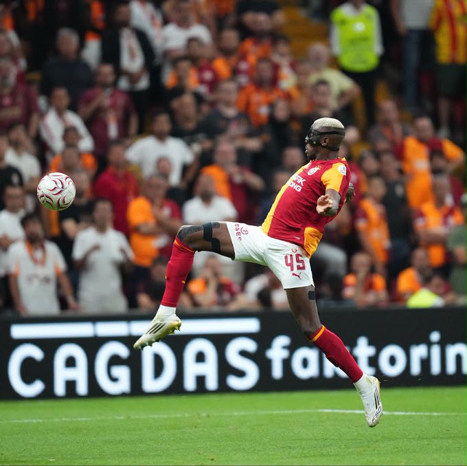 Victor Osimhen Trybal News Galatasaray player in red and yellow kit mid air kick during a match ball approaching from the left with cheering crowd in the stands behind trybalnews