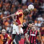 Two soccer players leap for a header as the ball comes down with a blurred stadium crowd behind them  trybalnews