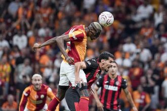 Two soccer players leap for a header as the ball comes down, with a blurred stadium crowd behind them.