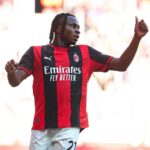 Soccer player in AC Milan red and black jersey celebrating with arm raised and thumbs up on the pitch  trybalnews