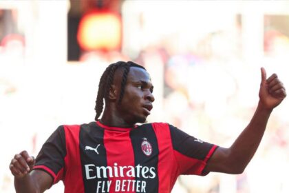 Soccer player in AC Milan red-and-black jersey celebrating with arm raised and thumbs up on the pitch.