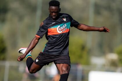 Soccer player in a black kit kicks a ball on a sunny training field.