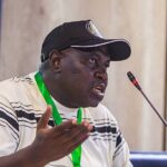 Man wearing a cap and striped shirt speaks at a panel gesturing with his left hand microphone in front  trybalnews