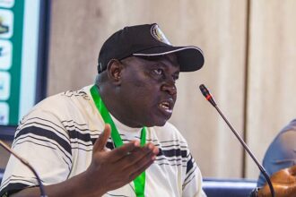 Man wearing a cap and striped shirt speaks at a panel, gesturing with his left hand, microphone in front.