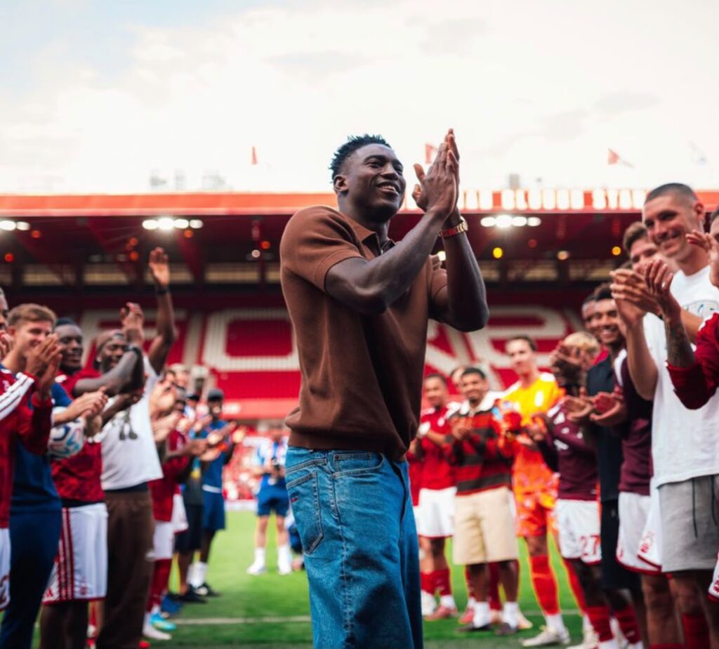 Young man in a brown shirt and jeans claps with teammates on a soccer field surrounded by fans and players applauding  trybalnews