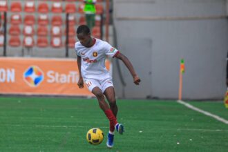Soccer player in a white kit with red trim dribbling a yellow ball on a green field during a match.
