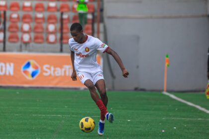 Soccer player in a white kit with red trim dribbling a yellow ball on a green field during a match.