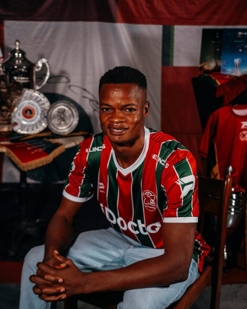 Musa Maliq Trybal News Smiling young man in a red and green striped soccer jersey sits indoors with trophies and banners visible in the background trybalnews