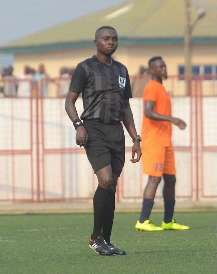 Plateau State referee Late Awang Solomon Maram Trybal News Soccer referee in a black kit standing on a green field with a blurred orange clad player in the background trybalnews