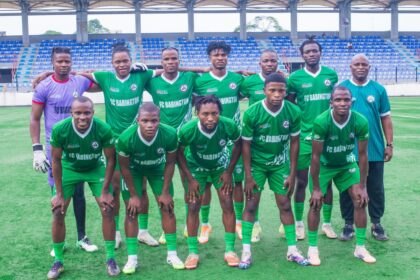 Group of soccer players in green uniforms posing for a team photo on a field with empty blue stadium seats in the background.