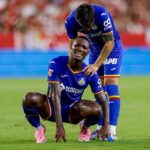 Two soccer players in blue uniforms on a green field one kneels in pain while a teammate stands beside him offering support  trybalnews