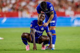 Two soccer players in blue uniforms on a green field; one kneels in pain while a teammate stands beside him offering support.