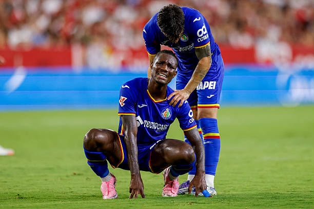 Two soccer players in blue uniforms on a green field one kneels in pain while a teammate stands beside him offering support  trybalnews