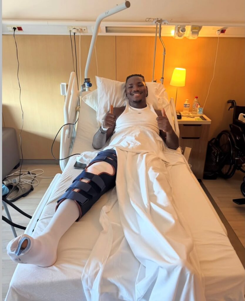 Christantus Uche Trybal News Smiling man in a hospital bed with a leg brace and bandages giving thumbs up hospital room with a nightstand and lamp in the background trybalnews