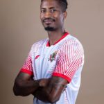 Portrait of a man with short dreadlocks wearing a white and red sports jersey arms crossed smiling slightly against a neutral backdrop looking at the camera  trybalnews