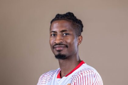 Portrait of a man with short dreadlocks wearing a white and red sports jersey, arms crossed, smiling slightly against a neutral backdrop, looking at the camera.