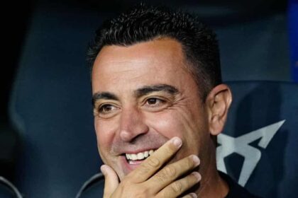 Smiling man with short dark hair, hand resting near his mouth in a dimly lit setting.