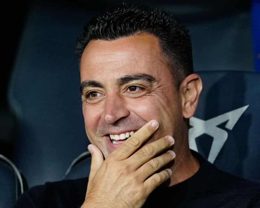 Smiling man with short dark hair hand resting near his mouth in a dimly lit setting  trybalnews