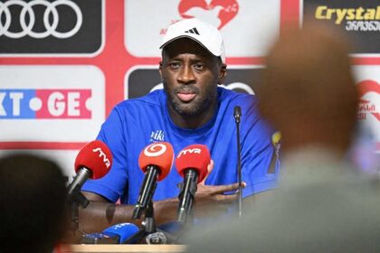 Yaya Toure, head coach of Slovan Bratislava, wearing a blue shirt and white Adidas cap, speaks at a press conference with microphones in front of him. The press conference followed the UEFA Champions League 2026/27 second qualifying round first leg match between Iberia Tbilisi and Slovan Bratislava at Mikheil Meskhi Stadium on July 21, 2026 in Tbilisi, Georgia.