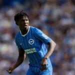 Medium shot of young football player Zadok Yohanna on the field during a match He is wearing the Brighton  Hove Albion home kit which features a blue jersey with thin white vertical stripes the club crest and the American Express sponsor logo His hair is styled in short twists and he is looking intently off camera to his left with a focused expression The stadium crowd behind him is heavily blurred  trybalnews