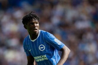 Medium shot of young football player Zadok Yohanna on the field during a match. He is wearing the Brighton & Hove Albion home kit, which features a blue jersey with thin white vertical stripes, the club crest, and the American Express sponsor logo. His hair is styled in short twists, and he is looking intently off-camera to his left with a focused expression. The stadium crowd behind him is heavily blurred.