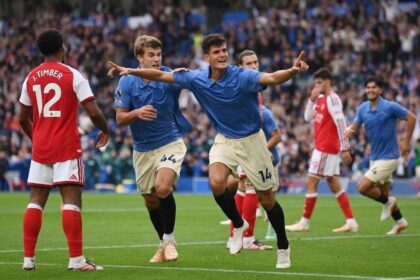 Brighton & Hove Albion midfielder Chema Andres, wearing a blue shirt and number 14 shorts, celebrates with teammate Luka Vuskovic after scoring, both with arms extended and pointing. Arsenal defender J. Timber in red and white number 12 kit stands nearby on the pitch at the American Express Stadium.