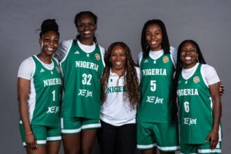 Nigeria’s women’s national basketball team, D’Tigress, pose together in green home jerseys with "NIGERIA" and "XE Jet" on the front. Players wear jersey numbers 1, 32, 5, and 6 and stand on either side of Head Coach Rena Wakama, who is in a white Nigeria polo and black pants. The team smiles in front of a plain grey backdrop.