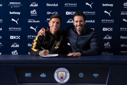 Enzo Fernandez signs his contract as a new Manchester City player at a signing table, wearing a black and gold training top. He is seated next to a Manchester City official with an arm around his shoulder. The backdrop features Manchester City and sponsor logos including Etihad, Puma, and Betway.