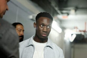 USMNT forward Folarin Balogun of the United States arrives at Fanatics Fest at Javits Center in New York, New York on July 17, 2026 ahead of a press conference for the FIFA World Cup 2026 Final. He wears a light blue striped shirt over a white t-shirt and a diamond earring, walking through a hallway with other people in the background.