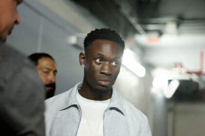 USMNT forward Folarin Balogun of the United States arrives at Fanatics Fest at Javits Center in New York, New York on July 17, 2026 ahead of a press conference for the FIFA World Cup 2026 Final. He wears a light blue striped shirt over a white t-shirt and a diamond earring, walking through a hallway with other people in the background.