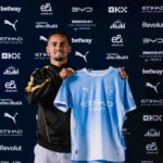 Iliman Ndiaye unveiled as a new Manchester City player smiling while holding up the clubs light blue home jersey with Etihad Airways on the front He wears a black and gold training jacket and stands in front of a backdrop with Manchester City and sponsor logos including Etihad Puma and Betway  trybalnews