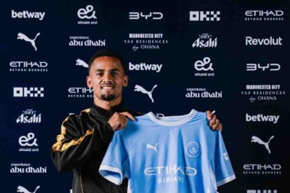 Iliman Ndiaye unveiled as a new Manchester City player, smiling while holding up the club’s light blue home jersey with "Etihad Airways" on the front. He wears a black and gold training jacket and stands in front of a backdrop with Manchester City and sponsor logos including Etihad, Puma, and Betway.