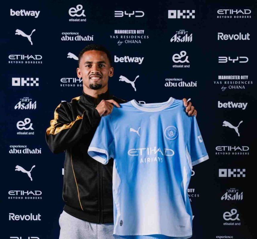 Iliman Ndiaye unveiled as a new Manchester City player, smiling while holding up the club’s light blue home jersey with "Etihad Airways" on the front. He wears a black and gold training jacket and stands in front of a backdrop with Manchester City and sponsor logos including Etihad, Puma, and Betway.