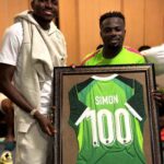 Nigerian footballers Victor Osimhen and Moses Simon smiling and posing together inside the Super Eagles dressing room while holding a large framed green Nigeria national team jersey The jersey has SIMON and the number 100 in white on the back commemorating Moses Simons 100th cap for Nigeria Osimhen on the left is wearing a white t shirt with a light grey hoodie draped over his shoulders and light grey shorts Simon on the right is wearing a bright neon green and pink Super Eagles training top The framed jersey has a black and gold ornate border Wooden lockers are visible in the background  trybalnews