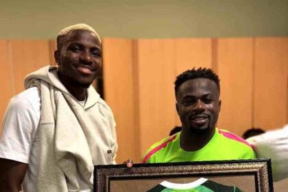 Nigerian footballers Victor Osimhen and Moses Simon smiling and posing together inside the Super Eagles dressing room while holding a large framed green Nigeria national team jersey. The jersey has "SIMON" and the number "100" in white on the back, commemorating Moses Simon's 100th cap for Nigeria. Osimhen, on the left, is wearing a white t-shirt with a light grey hoodie draped over his shoulders and light grey shorts. Simon, on the right, is wearing a bright neon green and pink Super Eagles training top. The framed jersey has a black and gold ornate border. Wooden lockers are visible in the background.