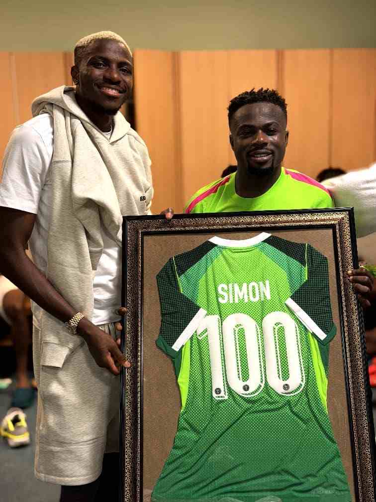 Nigerian footballers Victor Osimhen and Moses Simon smiling and posing together inside the Super Eagles dressing room while holding a large framed green Nigeria national team jersey The jersey has SIMON and the number 100 in white on the back commemorating Moses Simons 100th cap for Nigeria Osimhen on the left is wearing a white t shirt with a light grey hoodie draped over his shoulders and light grey shorts Simon on the right is wearing a bright neon green and pink Super Eagles training top The framed jersey has a black and gold ornate border Wooden lockers are visible in the background  trybalnews