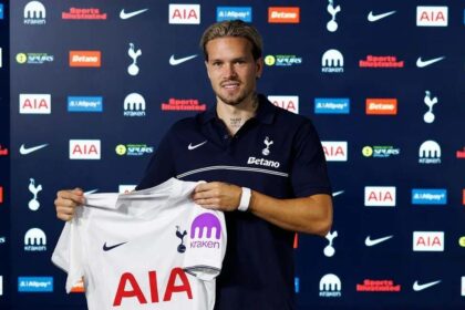 Mykhailo Mudryk unveiled as a new Tottenham Hotspur player after joining on loan from Chelsea. The Ukrainian winger smiles while holding up the club’s white home jersey with "AIA" and "Kraken" sponsors. He wears a navy blue Tottenham polo with "Betano" on the front and stands in front of a backdrop with Tottenham, Nike, AIA, and other sponsor logos.