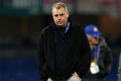 Chairman Todd Boehly of Chelsea FC walks across the pitch with his hands in his pockets after the UEFA Champions League match. He is wearing a black zip-up jacket over a black shirt, light-colored pants, and a UEFA accreditation lanyard. The stadium background is blurred.
