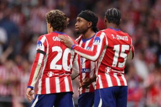 Ademola Lookman of Atletico Madrid, wearing a black headband and red-and-white striped Atletico jersey, celebrates with teammates Giuliano Simeone (number 20) and Jonathan David (number 15) after scoring his team's second goal during the UEFA Champions League 2026/27 League Phase MD2 match between Atletico de Madrid and Manchester United at Estadio Metropolitano on October 13, 2026, in Madrid, Spain.