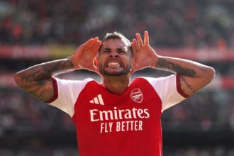 Arsenal midfielder Bruno Guimaraes celebrates with hands cupped around his ears after scoring during a Premier League match at Emirates Stadium. He is wearing the red and white Arsenal home kit with visible arm tattoos.