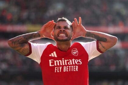 Arsenal midfielder Bruno Guimaraes celebrates with hands cupped around his ears after scoring during a Premier League match at Emirates Stadium. He is wearing the red and white Arsenal home kit with visible arm tattoos.