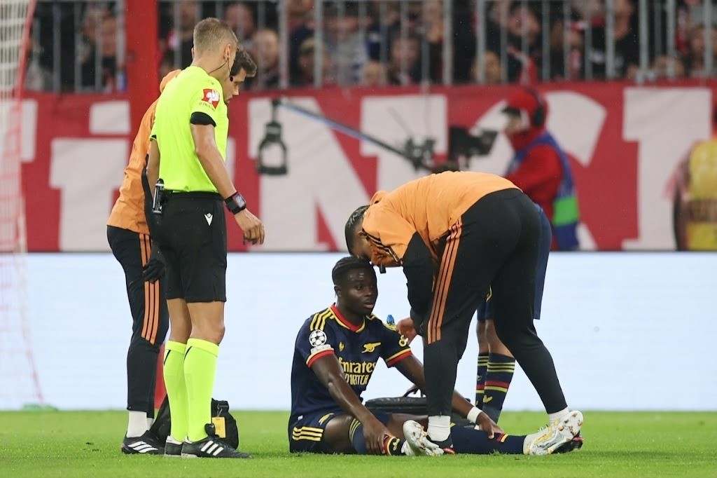 Bukayo Saka Trybal News Arsenal winger Bukayo Saka in navy away kit sitting on grass holding his leg while Arsenal physio in orange jacket attends to him with referee in yellow kit standing nearby at Football Arena Munich trybalnews