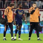 Arsenal winger Bukayo Saka in navy away kit walking off pitch supported by two Arsenal medical staff in orange jackets at Football Arena Munich after quadriceps injury  trybalnews