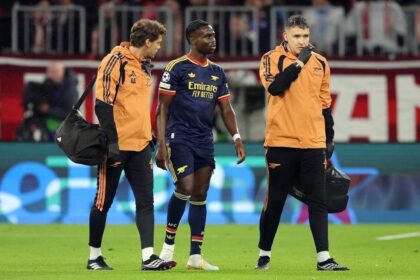 Arsenal winger Bukayo Saka in navy away kit walking off pitch supported by two Arsenal medical staff in orange jackets at Football Arena Munich after quadriceps injury.