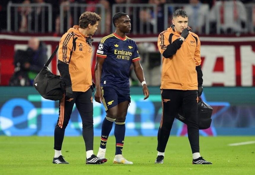 Arsenal winger Bukayo Saka in navy away kit walking off pitch supported by two Arsenal medical staff in orange jackets at Football Arena Munich after quadriceps injury  trybalnews