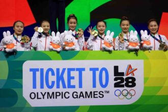 Six Chinese female gymnasts in white tracksuits holding gold medals and white bunny mascots in orange shirts smile on podium behind sign reading Ticket To LA 28 Olympic Games with Olympic rings logo  trybalnews