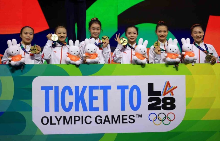 Six Chinese female gymnasts in white tracksuits holding gold medals and white bunny mascots in orange shirts smile on podium behind sign reading Ticket To LA 28 Olympic Games with Olympic rings logo  trybalnews