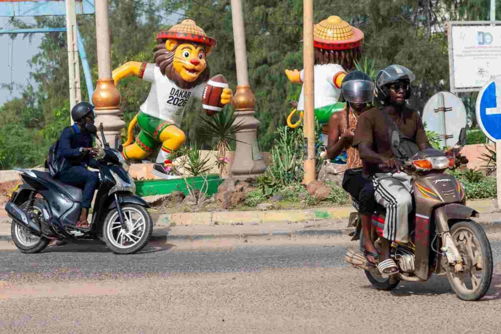 Colorful lion mascot Ayo wearing Dakar 2026 t shirt and holding rugby ball on roundabout island with two motorcycles riding past on road  trybalnews