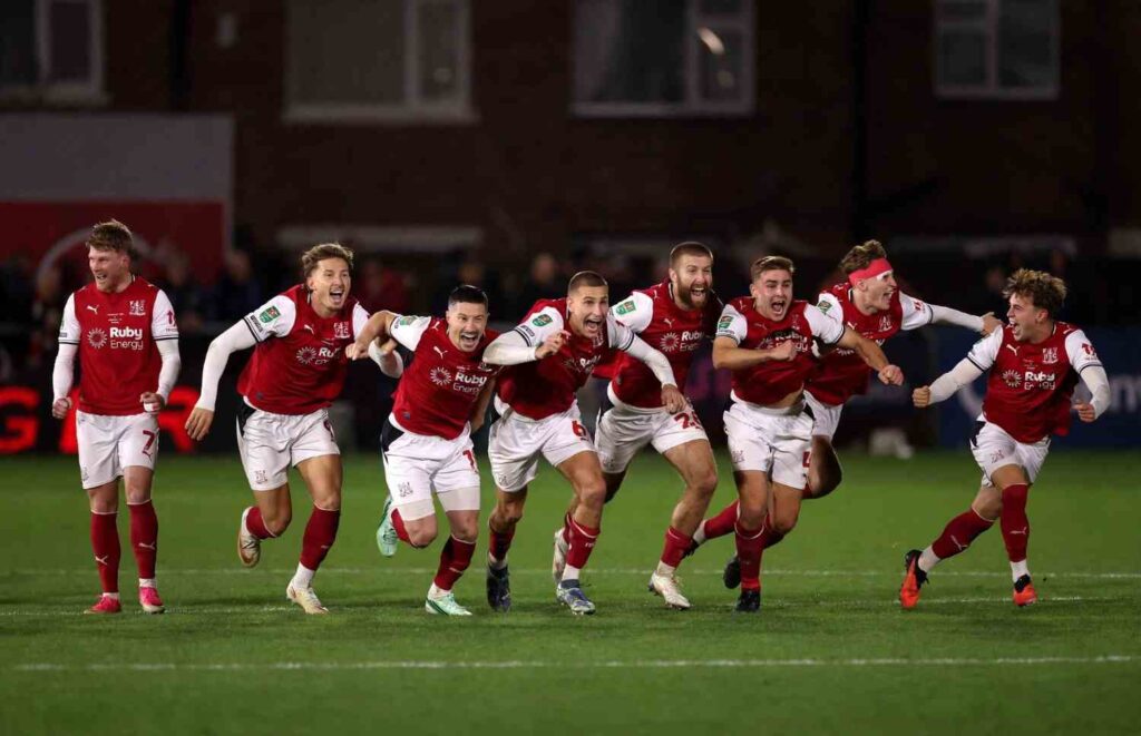 Eight Fleetwood Town players in red and white kits running and shouting in celebration on pitch at night  trybalnews