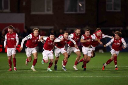 Eight Fleetwood Town players in red and white kits running and shouting in celebration on pitch at night